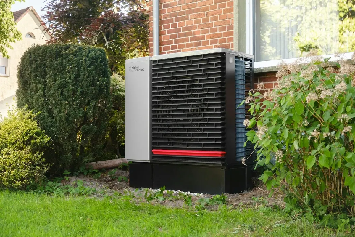 An outdoor heat-pump unit beside a brick house and garden.
