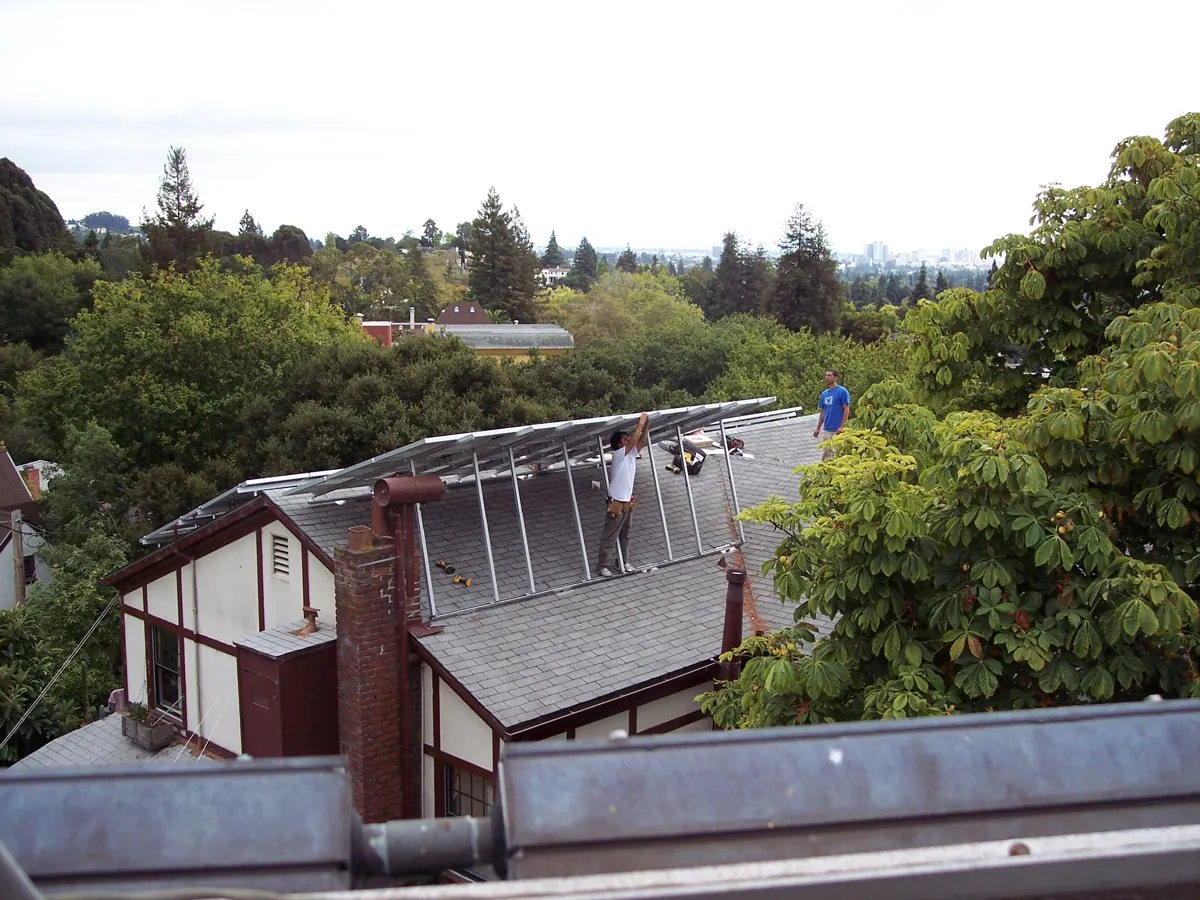 Installers work beneath tilted photovoltaic panels on a sloped house roof.