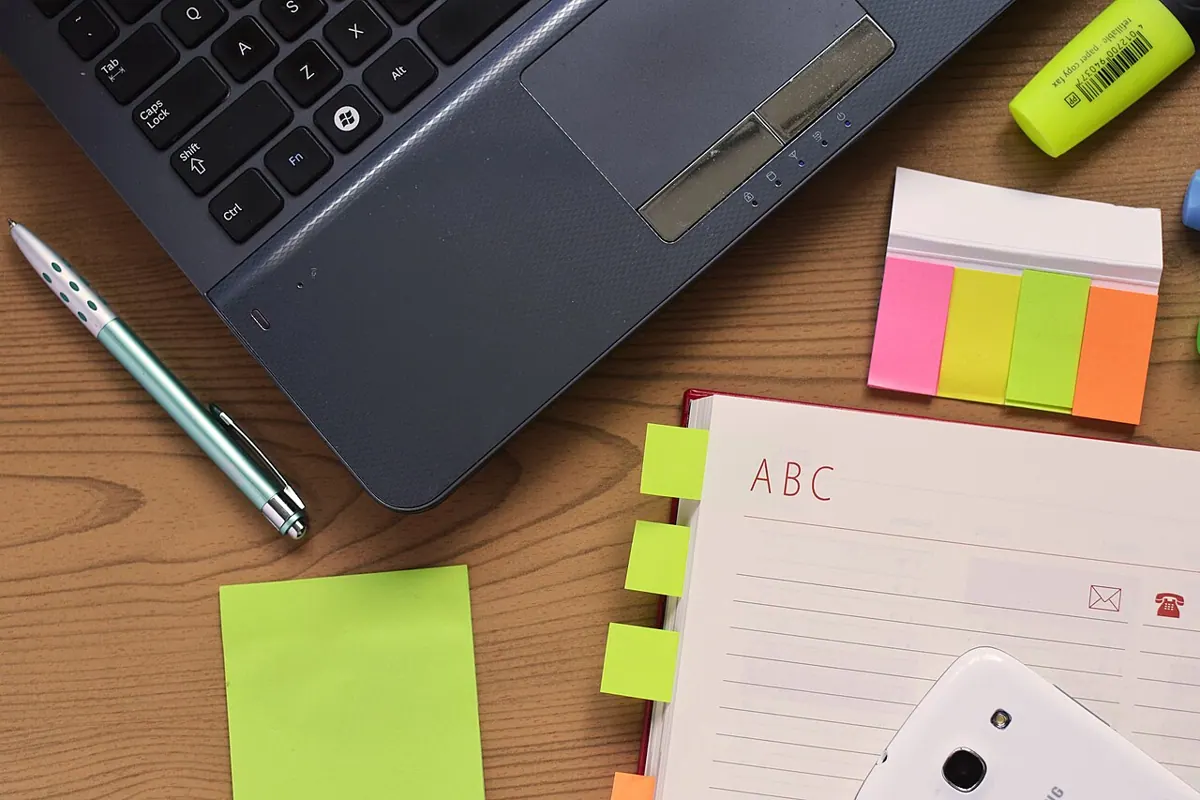 A laptop, notepad, pen and colorful sticky notes arranged on a wooden desk.